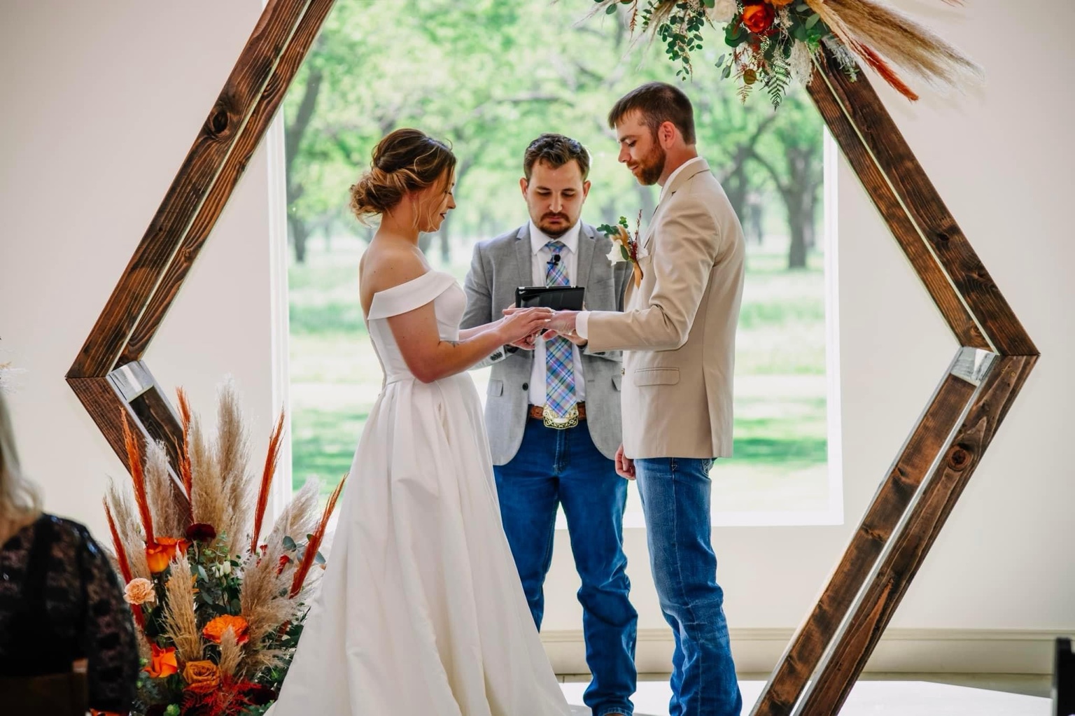 A couple exchanging rings with Eric beneath a wooden hexagon ceremony arch
