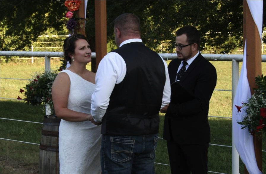 A couple sharing their vows with Eric beneath a white draped outdoor wedding arch